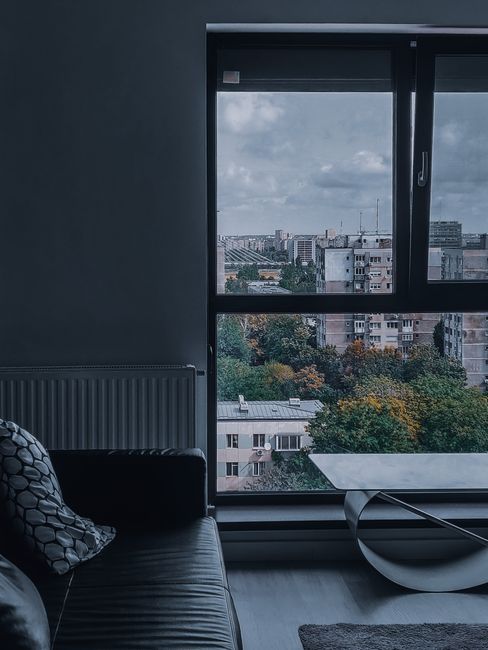 Indoor scene showing a modern living room with a large window providing a view of a cityscape with multi-story residential and commercial buildings, and trees with autumn foliage. The room features a 