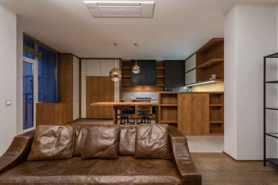 Indoor scene showing a modern living room with a large window providing a view of a cityscape with multi-story residential and commercial buildings, and trees with autumn foliage. The room features a 